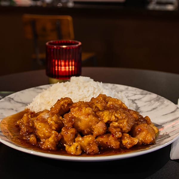 Orange chicken with white rice on a marble plate, with chopsticks and a menu on a dark table.