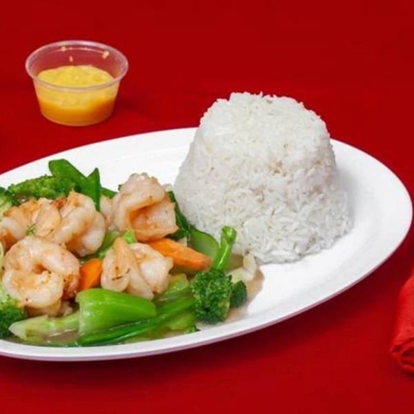 Plate of shrimp stir-fried with mixed vegetables and white rice, served with two dipping sauces on a red tablecloth.