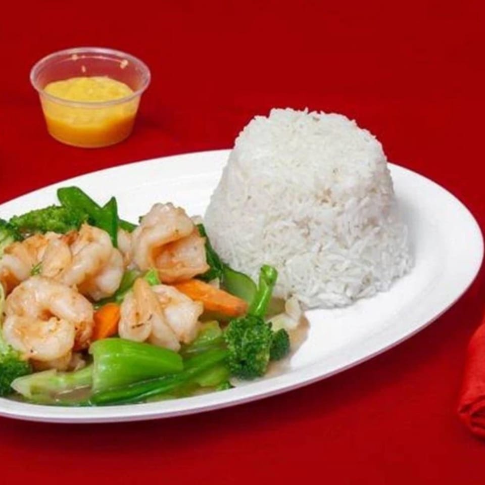 Plate of shrimp stir-fried with mixed vegetables and white rice, served with two dipping sauces on a red tablecloth.