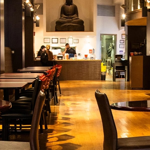 Restaurant interior with dark wood tables, red chairs, a Buddha statue, and staff behind a counter.