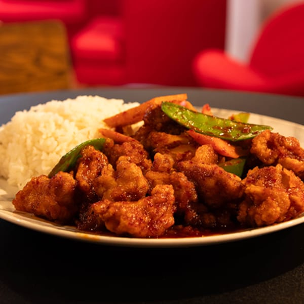 Sweet and sour chicken with rice and vegetables on a table with a candle and red chairs.
