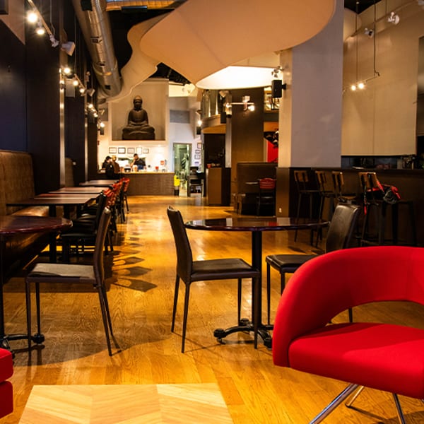 Spacious restaurant interior with wooden floors, modern tables, red chairs, and a Buddha statue in the background.